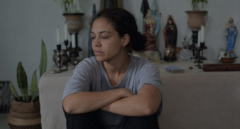 3 sinais de que você está em burnout espiritual e o que fazer 1 Mulher de camiseta cinza sentada no chão diante de altar com velas apagadas e baralho fechado, expressão de vazio e indiferença, representando o primeiro sinal do burnout espiritual.