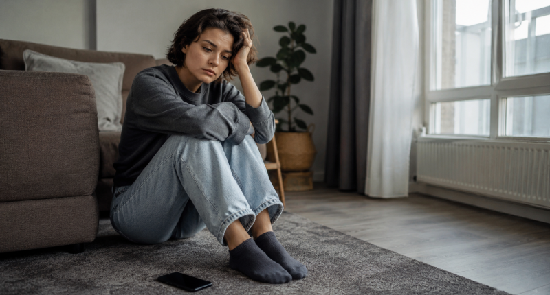 Mulher sentada no chão com celular virado para baixo ao lado, expressão vazia e frustrada, representando o padrão de busca de validação externa que afasta o amor verdadeiro.