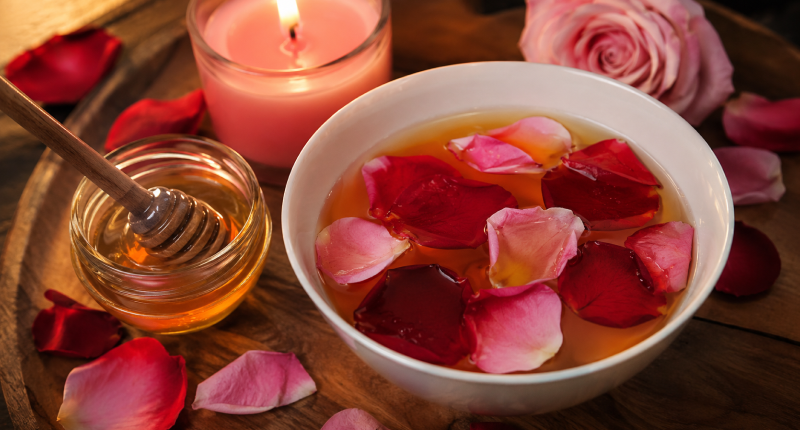 Tigela com pétalas de rosa vermelha e cor-de-rosa em água de mel com vela cor-de-rosa acesa ao fundo, representando os ingredientes do banho de atração para despertar o magnetismo amoroso.