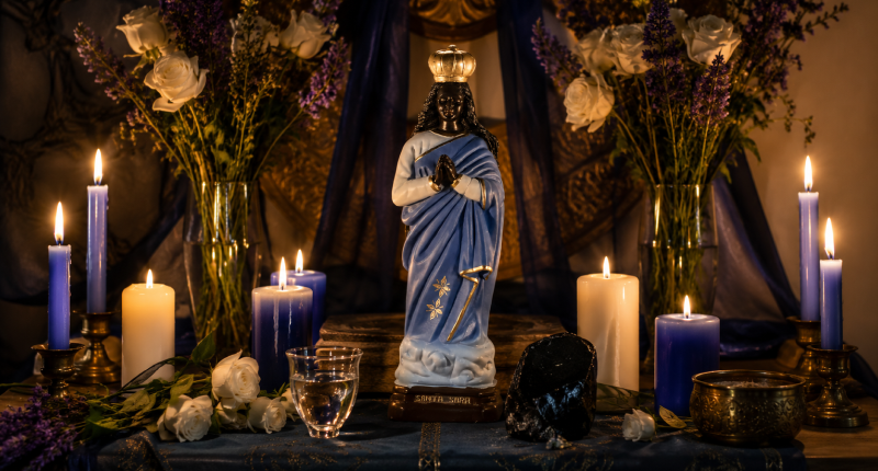Altar de Santa Sara Kali com estatueta, velas azuis e brancas acesas, flores frescas, água e obsidiana sobre tecido azul, representando o espaço sagrado de conexão com a grande mãe espiritual da ancestralidade.