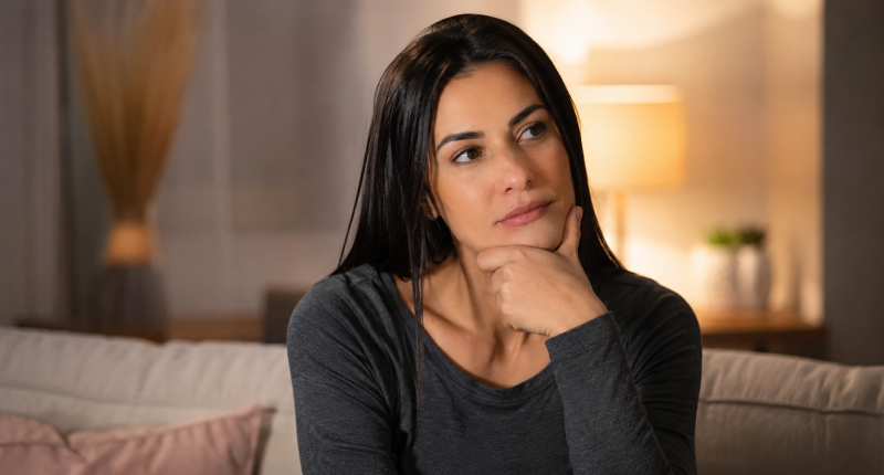 Mulher de blusa cinza sentada no sofá com mão no queixo e expressão pensativa e intrigada, representando a introspecção de quem carrega a pergunta sobre a própria mediunidade sem ainda ter uma resposta.